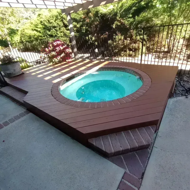 Small round turquoise hot tub on a wooden deck in a fenced backyard, with greenery around it