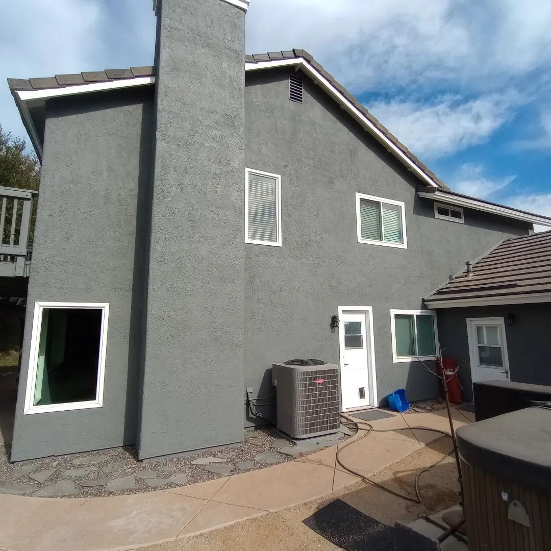 Gray stucco house exterior with patio, windows, door, and air-conditioning unit under a blue sky