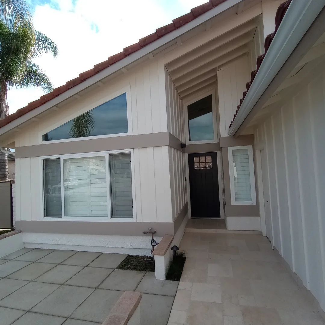 Front entrance of a beige house with large windows, tiled walkway, and open doorway under a sloped roof