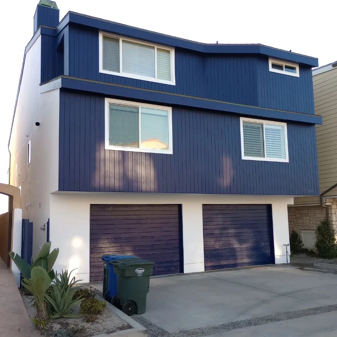 Blue multi-story house with white garage doors, driveway, and a green trash bin at the curb