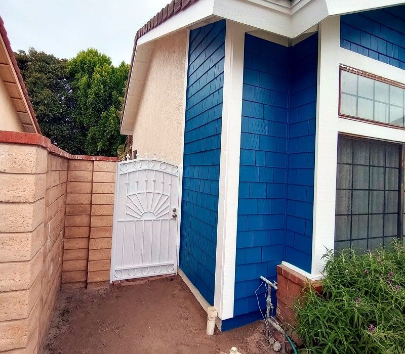 Narrow side yard beside a blue house with a white gate and tan brick wall