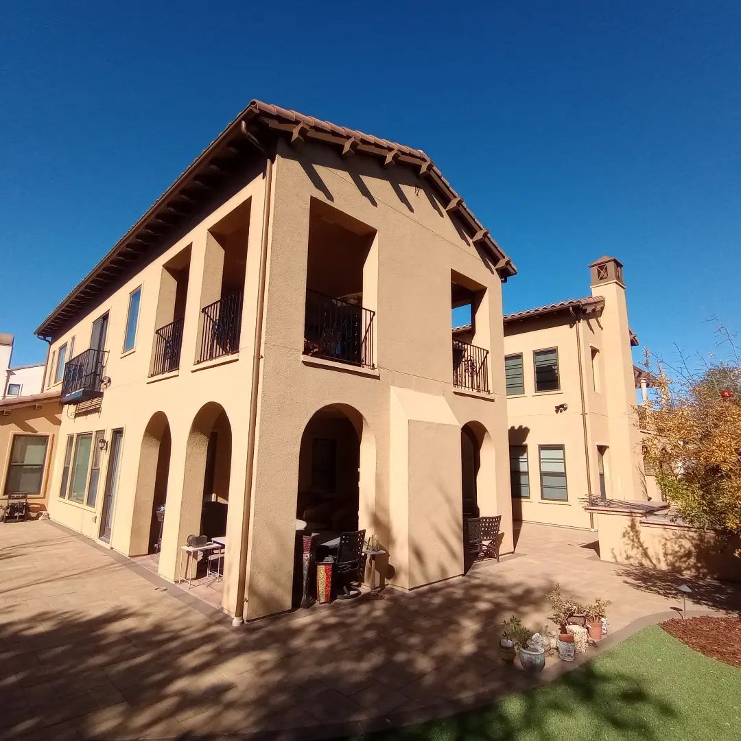 Tan stucco two-story house with arched porch and terracotta roof in bright sunlight
