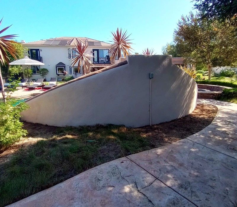 Outdoor garden with a curved concrete wall and walkway in front of a white house.