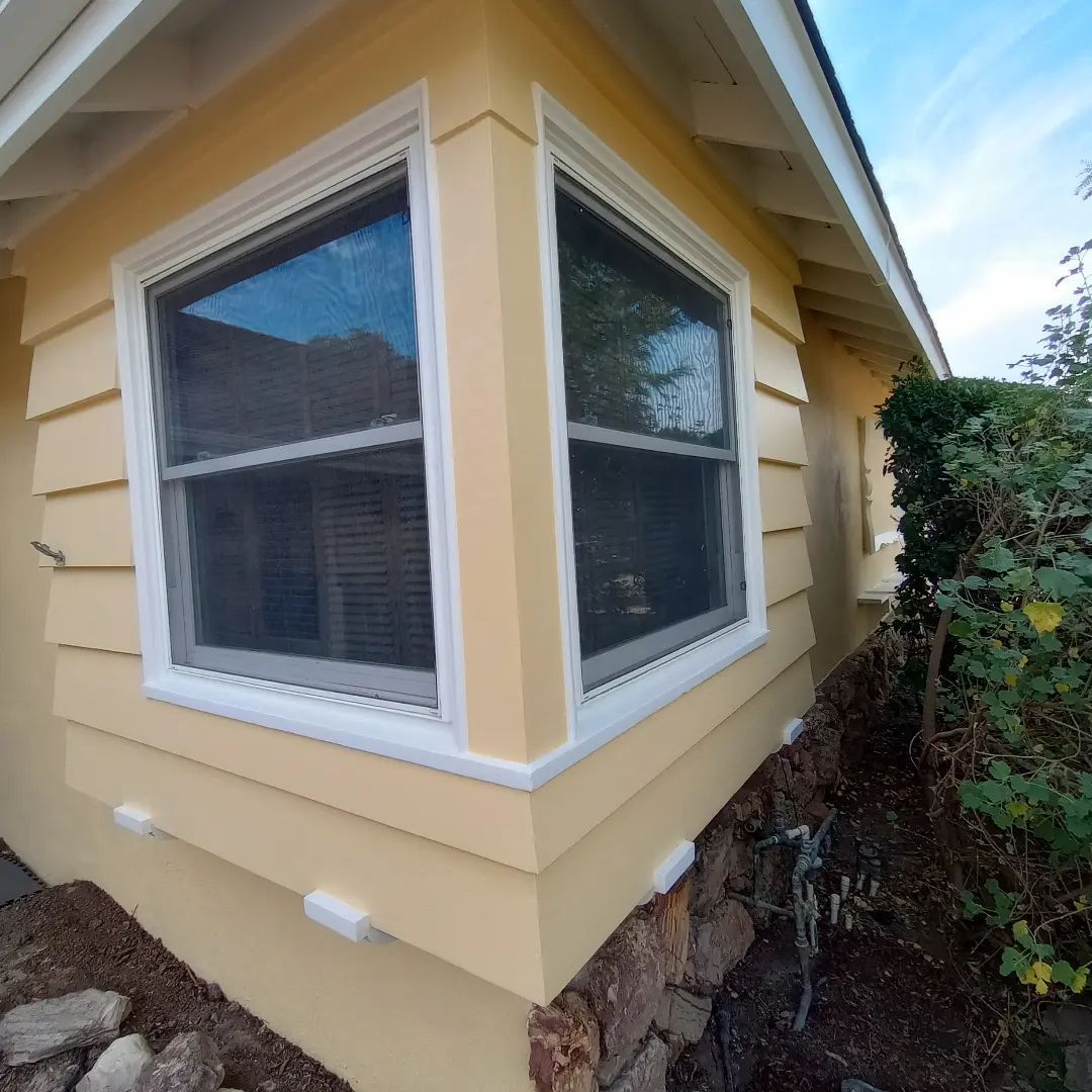 Corner of a yellow house with white-trimmed windows and stone foundation, beside shrubs and sky