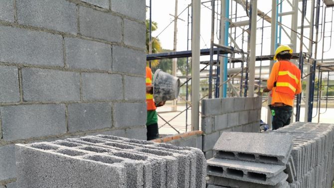 Workers in orange safety vests building a cinder block wall at a construction site.