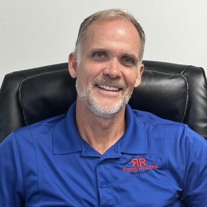 Man in blue polo shirt smiles, seated in a black chair; logo on shirt reads