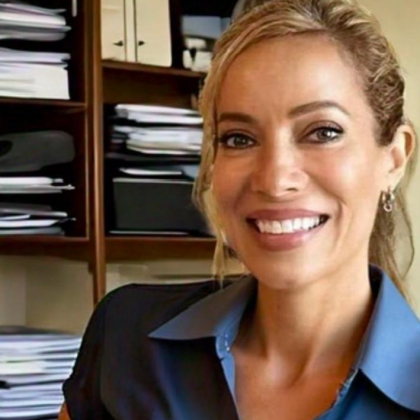 Woman smiles in front of a shelf with papers. She wears a blue shirt; natural light.