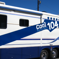 White and blue Cool 104.7 radio station trailer with windows and twin wheels, parked outdoors