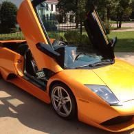 Orange sports car with scissor doors open, parked outdoors on a sunny day.