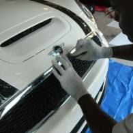 Hands install a chrome emblem on a white car grille with a tool.