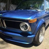 Blue vintage car parked outdoors, front-left view, with chrome wheel and round headlights.