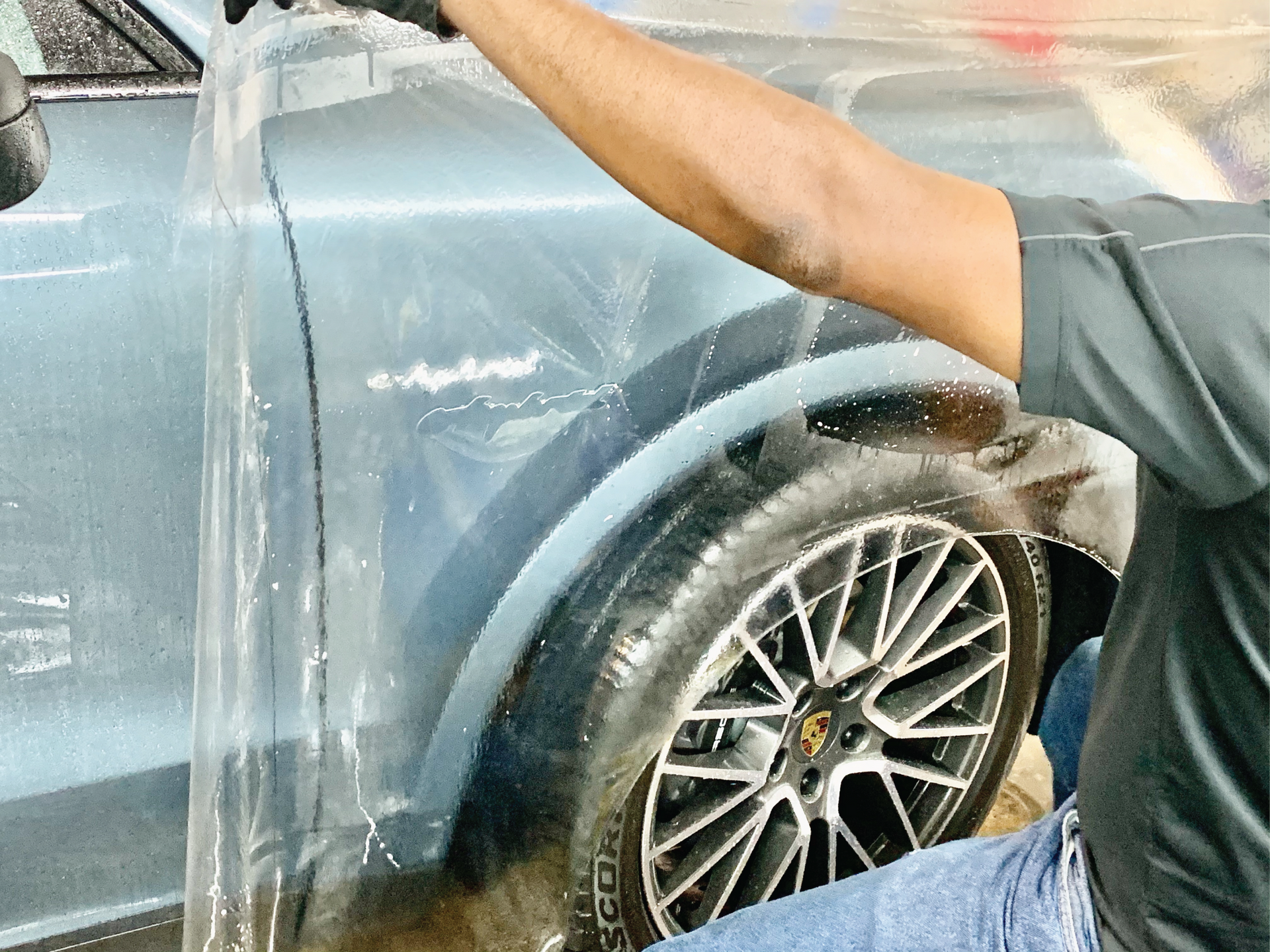 Person spraying water on a blue car’s rear wheel and side panel during a wash