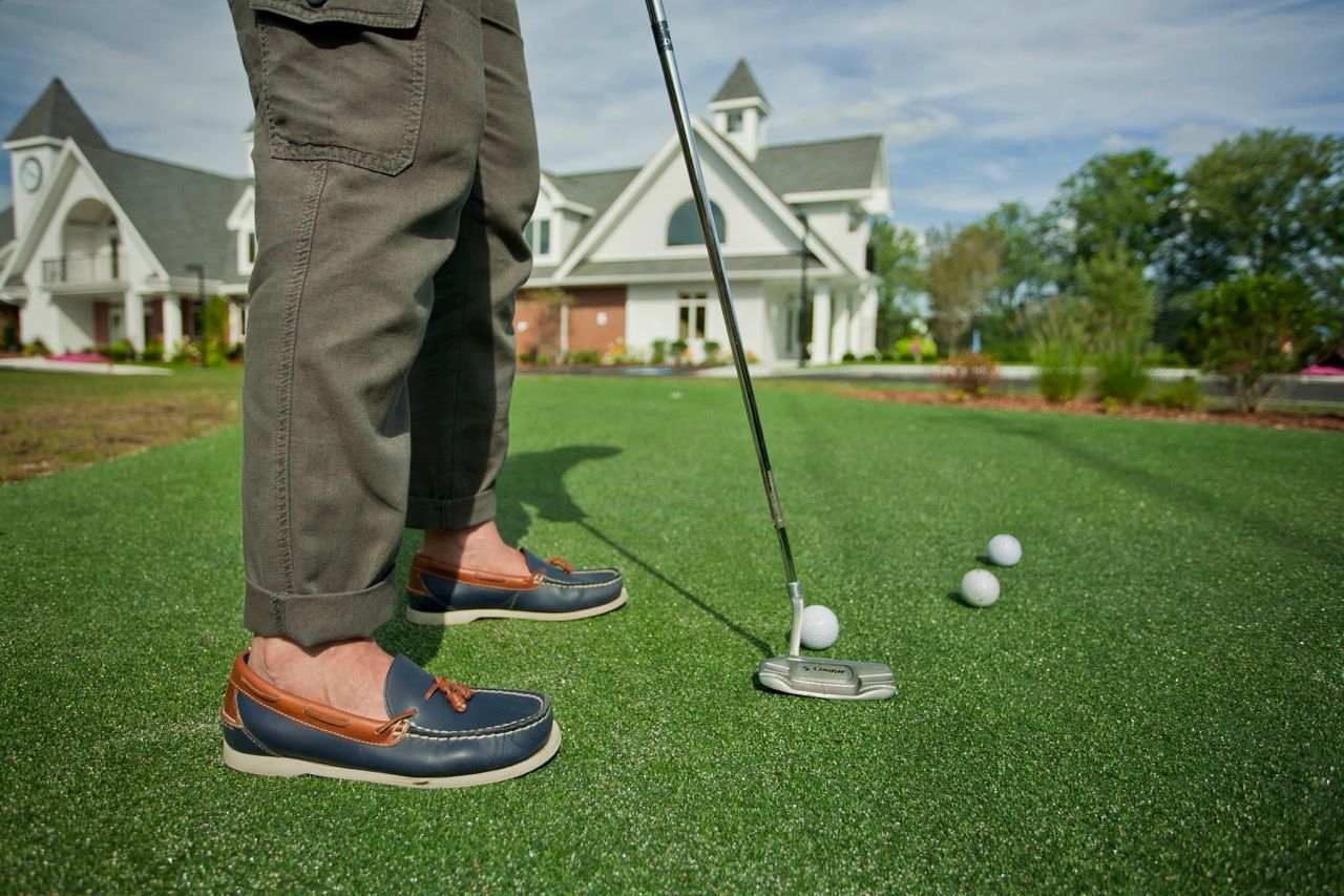 Person in navy loafers standing with a putter on a putting green with golf balls at a residential community.