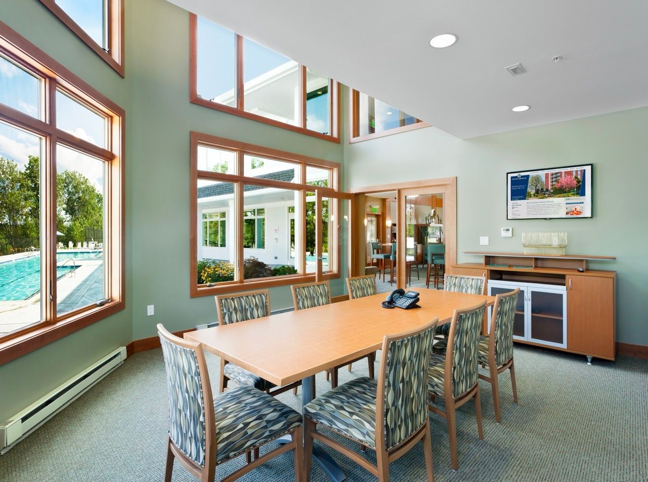 Interior community lounge with a long table, upholstered chairs, and windows overlooking the pool.