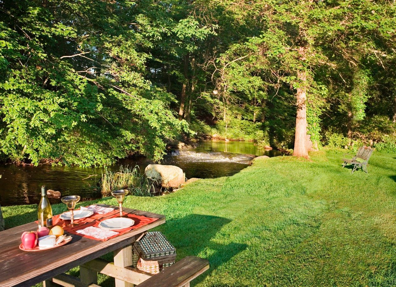 Picnic table with wine and plates set by a creek, surrounded by green lawn and trees.