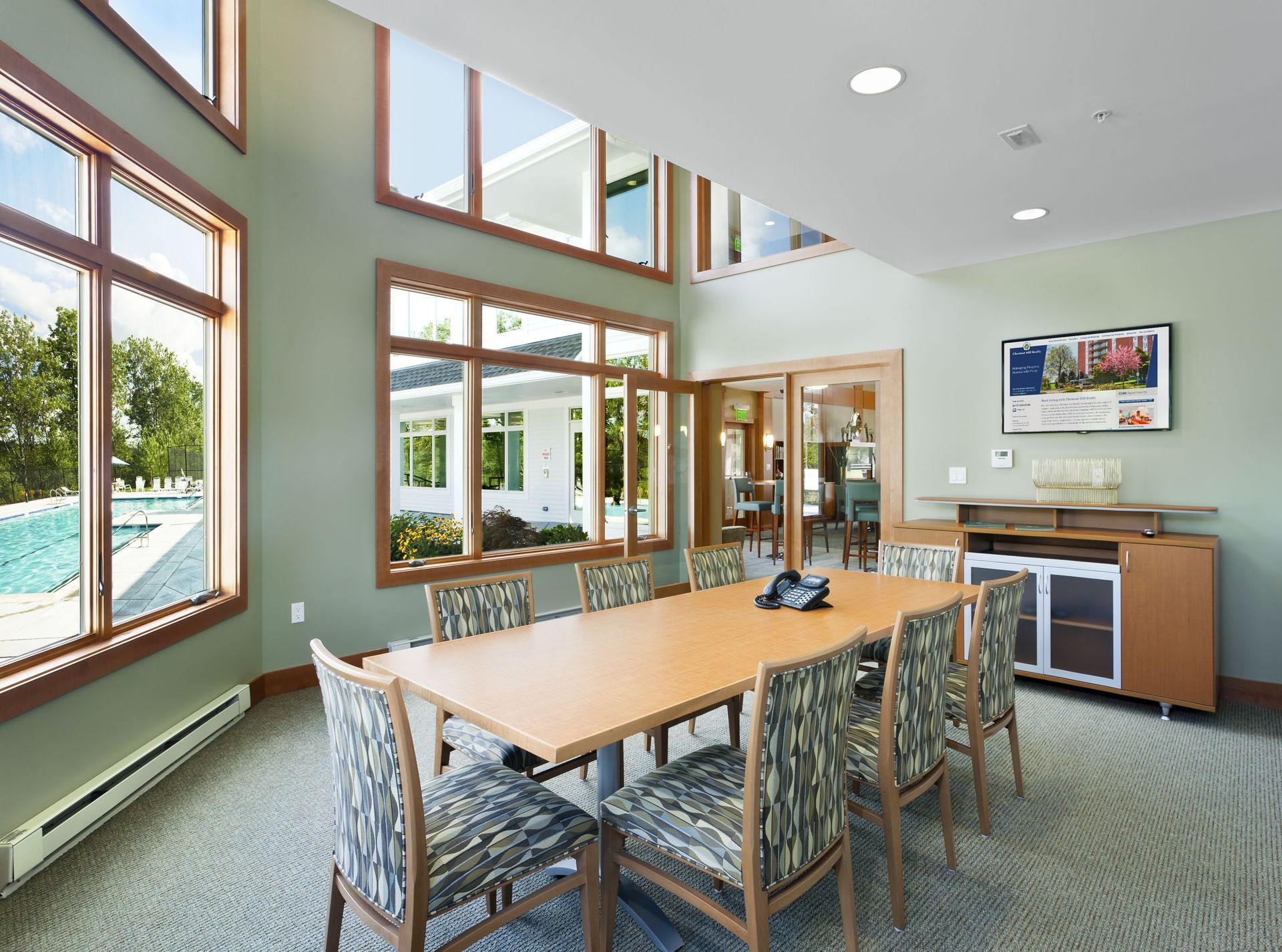 A bright, modern conference room with a large wooden table and six patterned chairs, set against tall windows.