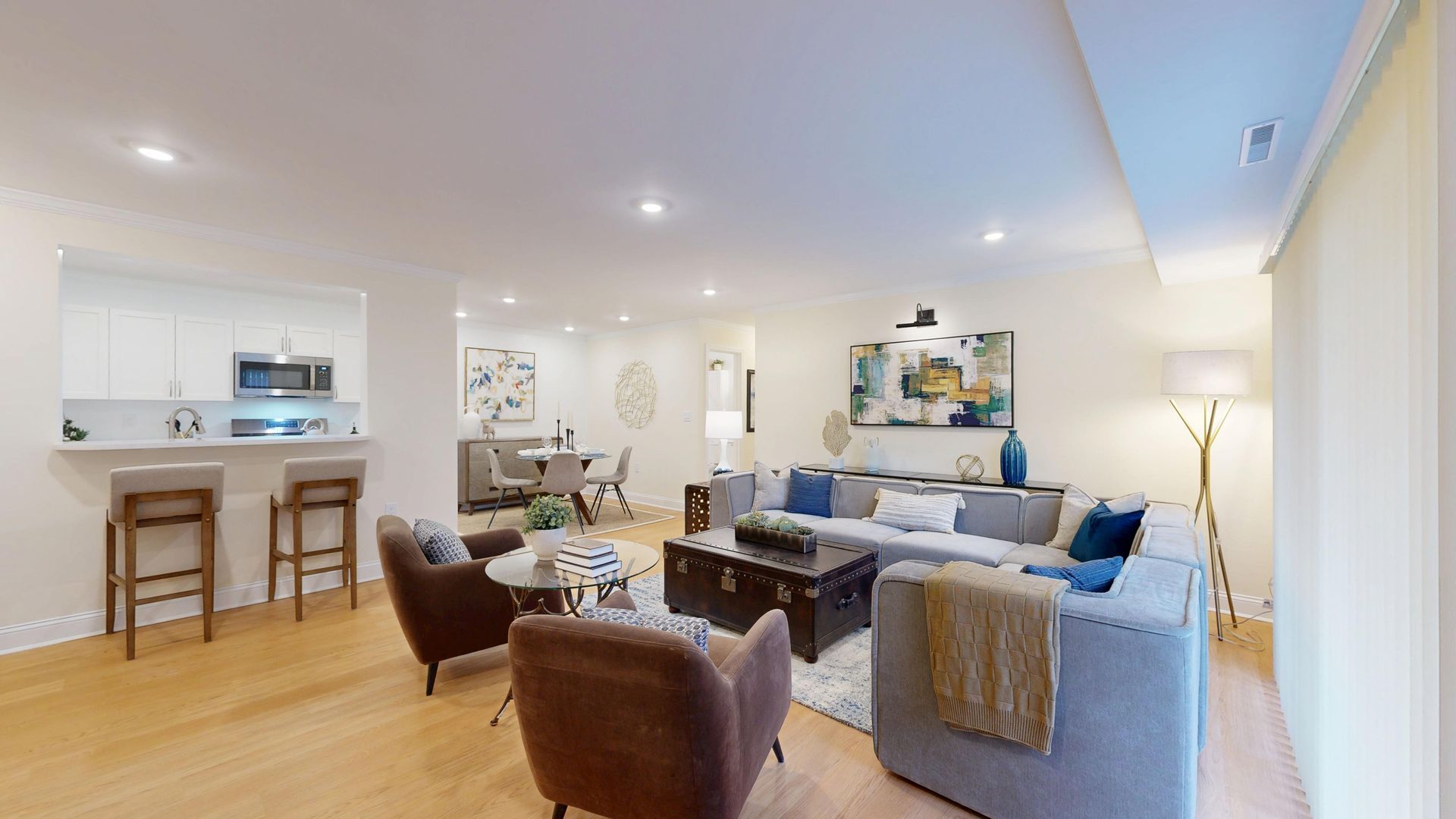 Bright open-concept living room with gray sectional, brown chairs, kitchen bar stools, and light wood floors