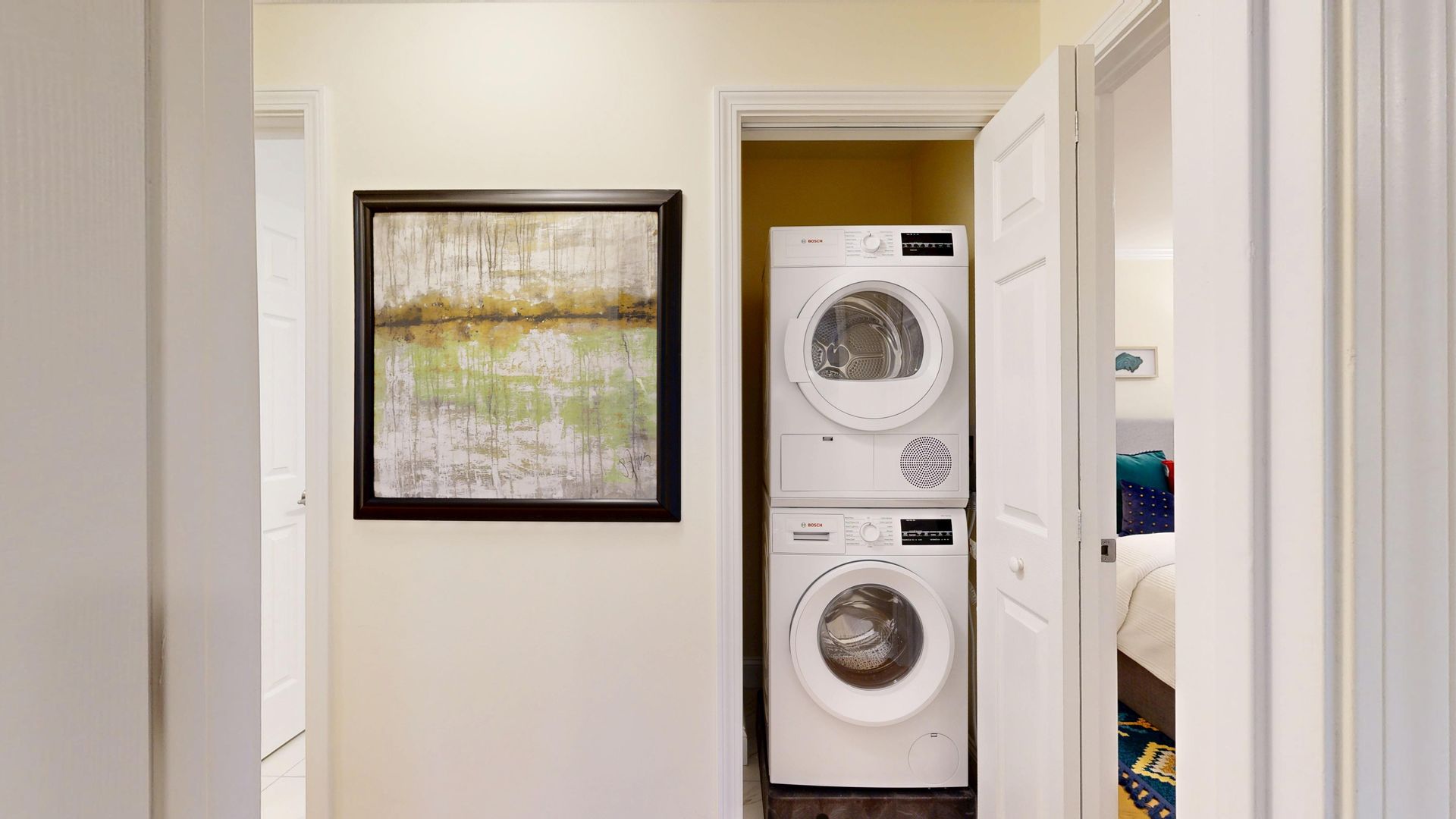 Hallway view of stacked white washer and dryer beside a framed landscape print.