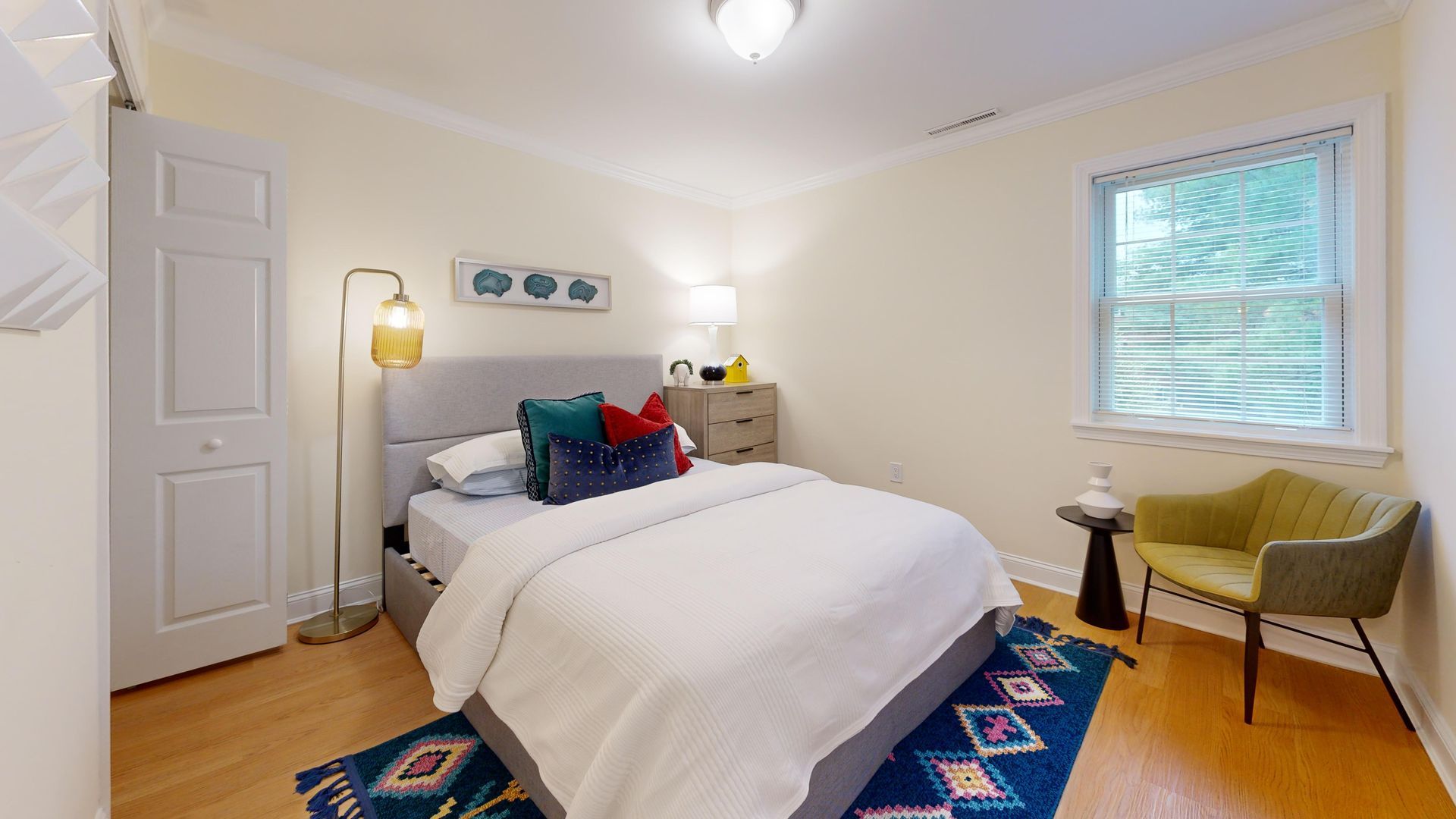 Bright bedroom with a made bed, white walls, blue rug, floor lamp, and window with sheer curtains.