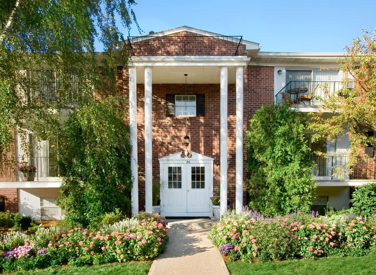 Exterior entrance of a brick apartment building with white doors, columns, and landscaped flowers.