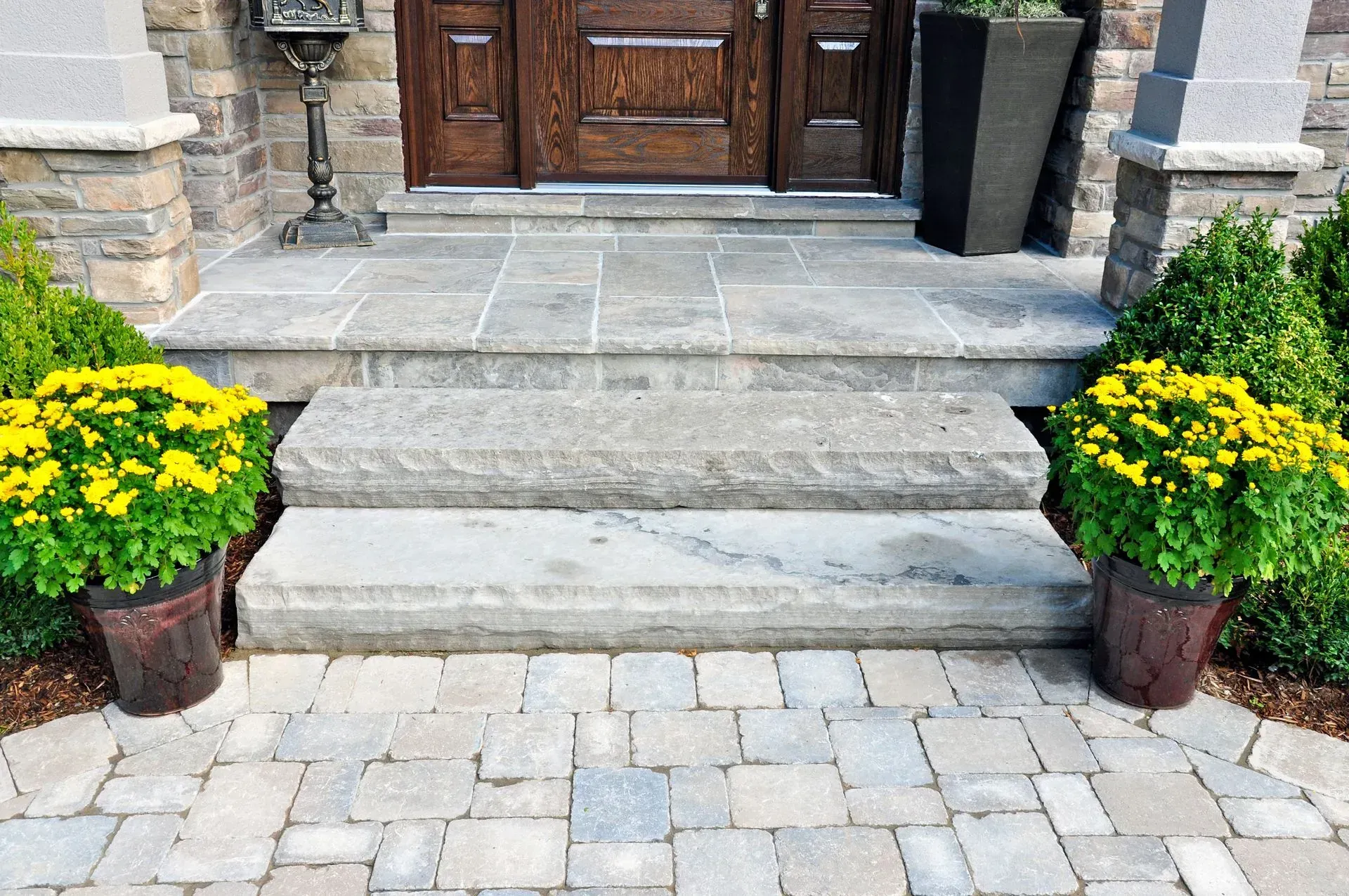 Stone front steps leading to a wooden door, flanked by potted yellow flowers and brick columns.