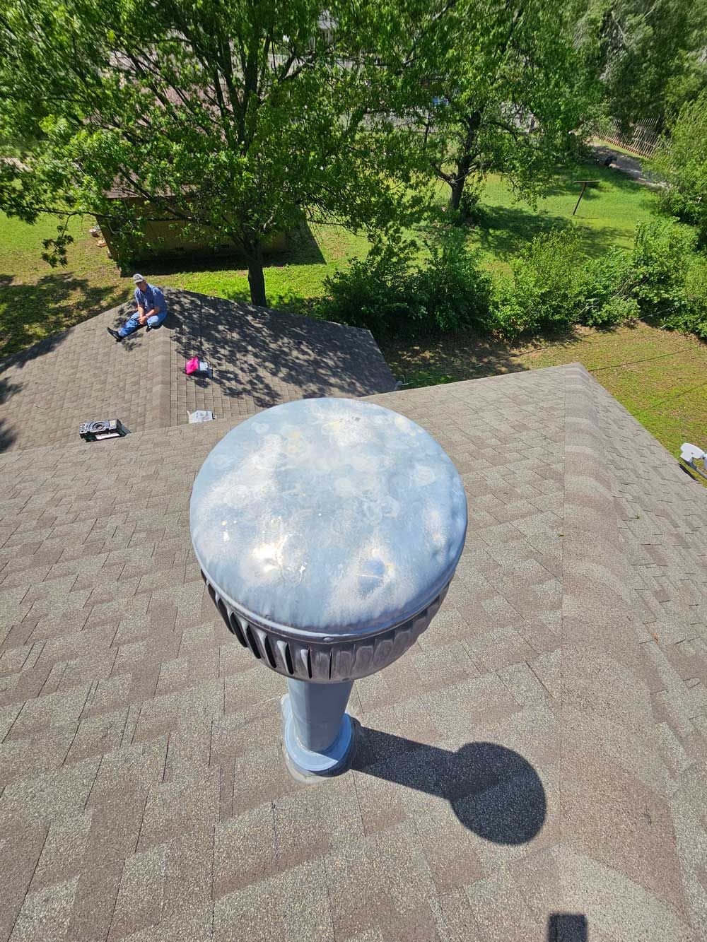 Blue roof vent on a brown shingle roof with green trees and grass in the background.