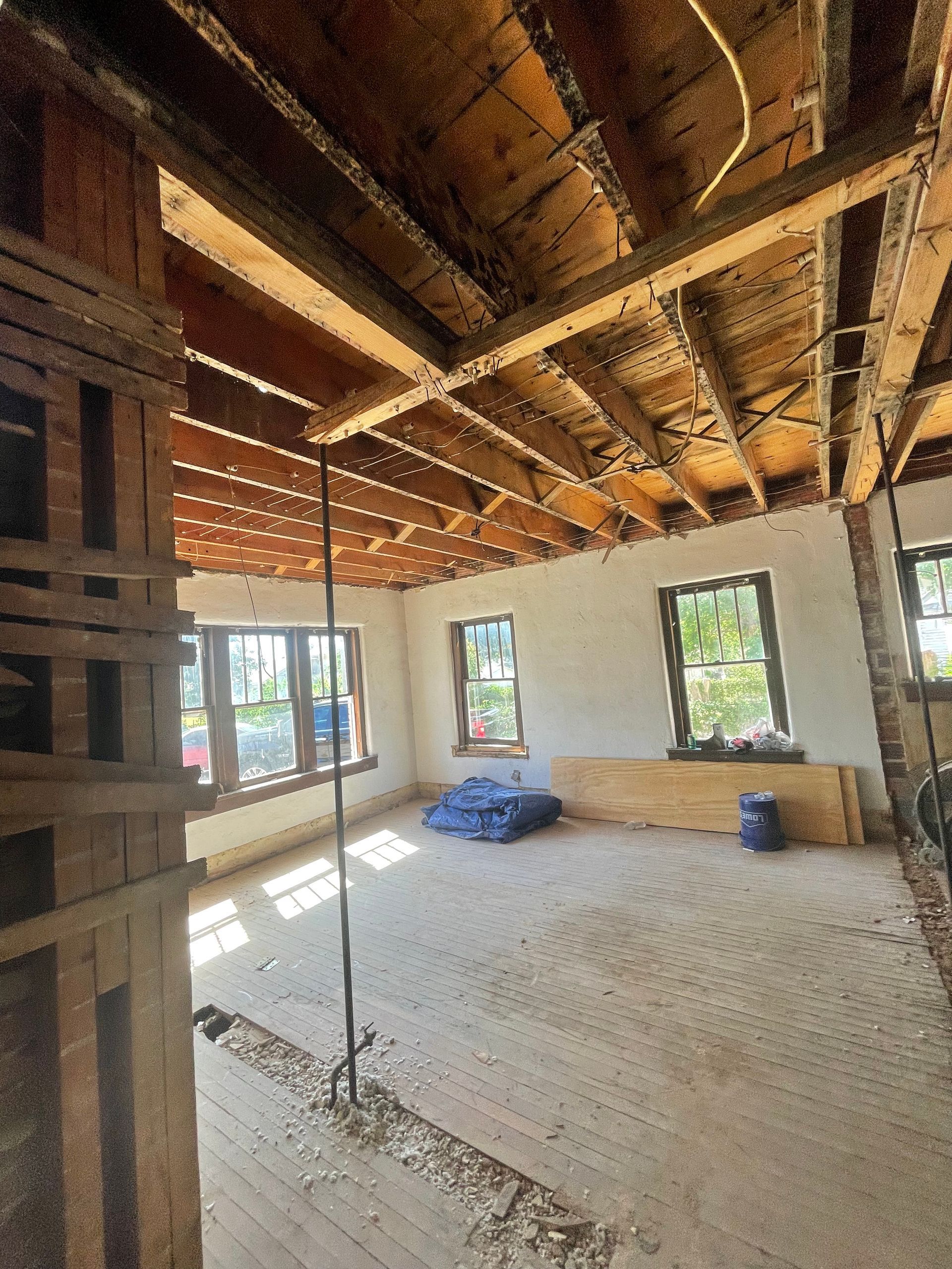 Room under construction: exposed ceiling beams, bare floor, windows, materials, and a support column.