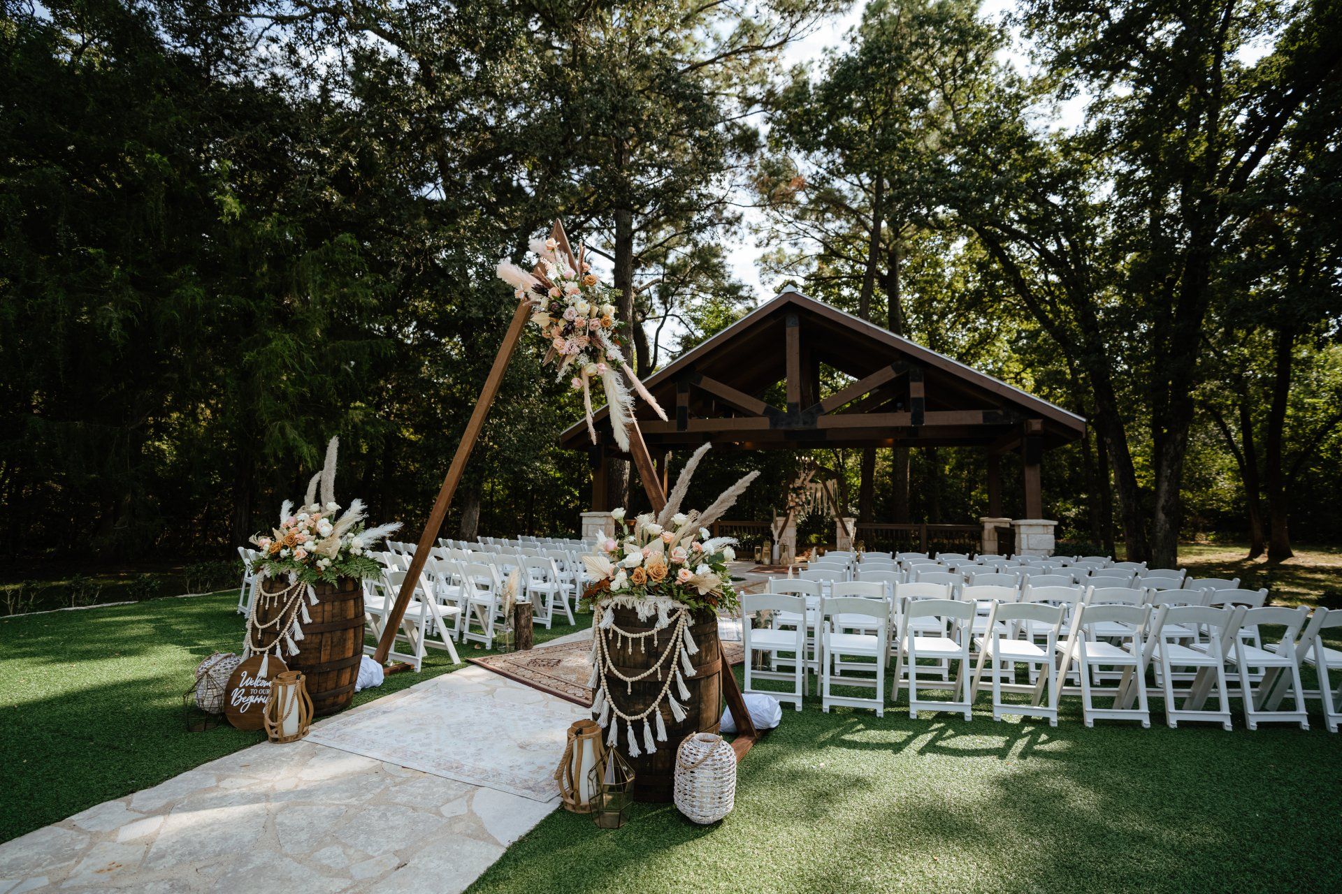 An outdoor wedding ceremony setup with rows of white chairs facing a wooden archway decorated with dried flowers and barrels.