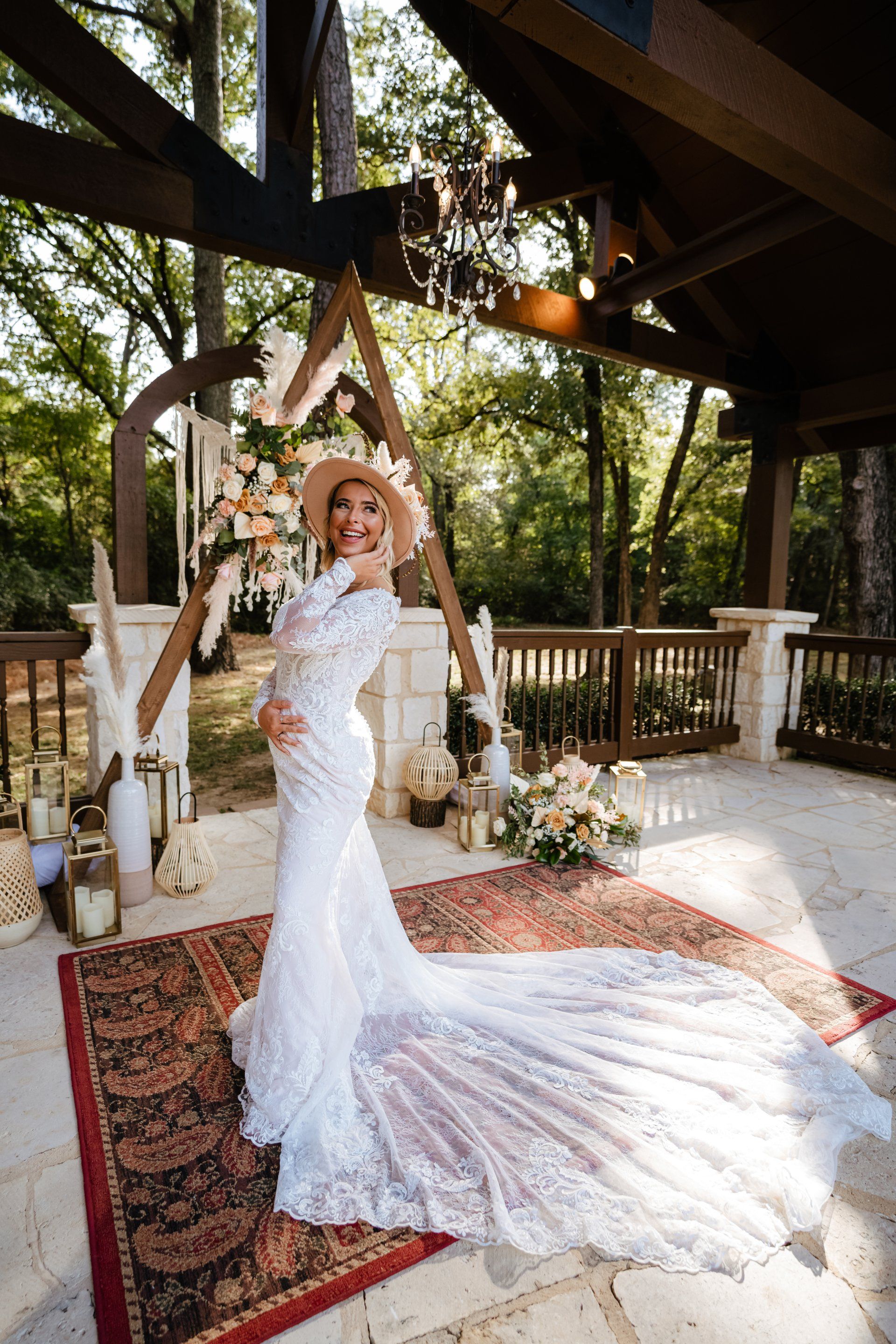 A person in a lace wedding dress and hat poses on a patterned rug under a wooden pavilion with floral decorations.