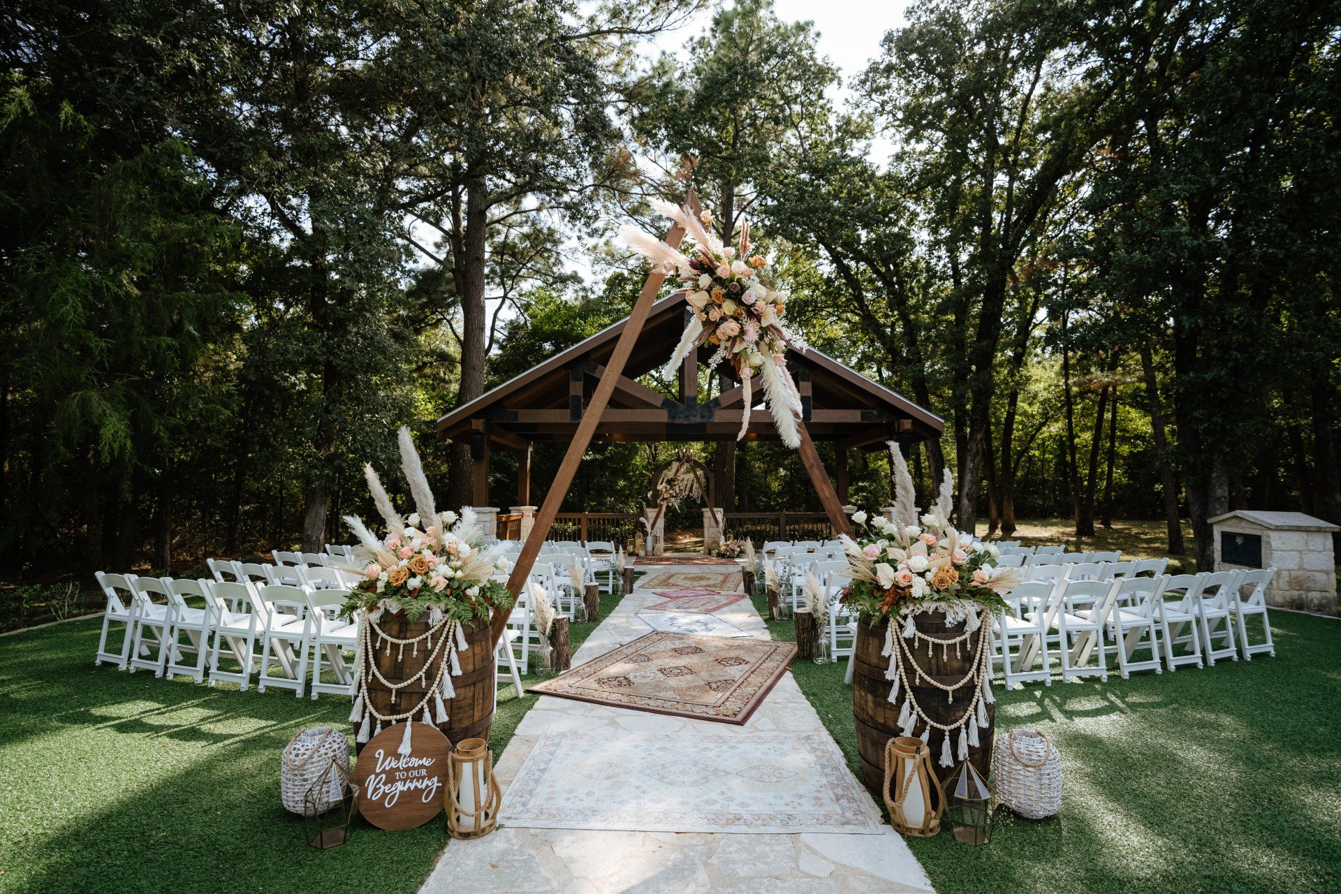 Outdoor wedding ceremony setup with white chairs, a triangular wooden arch, and floral arrangements on barrels.