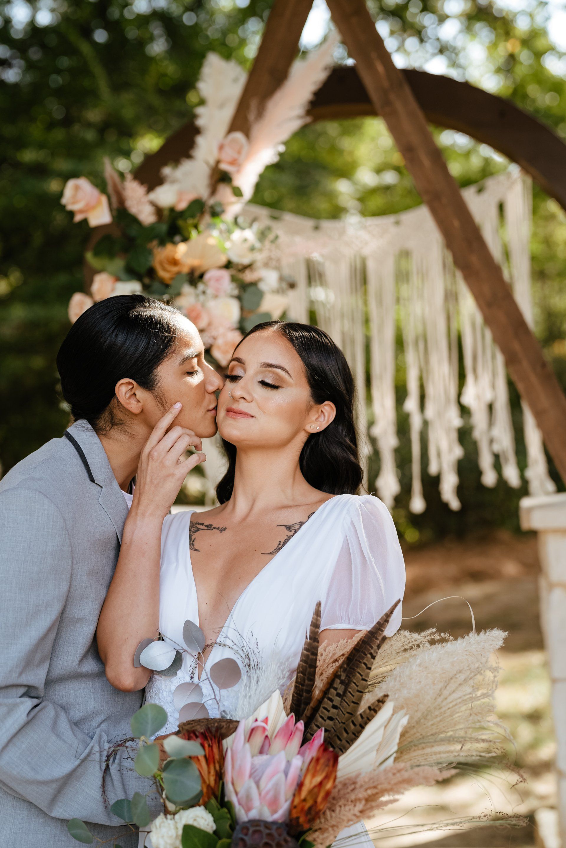 A person in a suit kisses the cheek of a person in a white wedding dress before a floral arch and macrame backdrop.