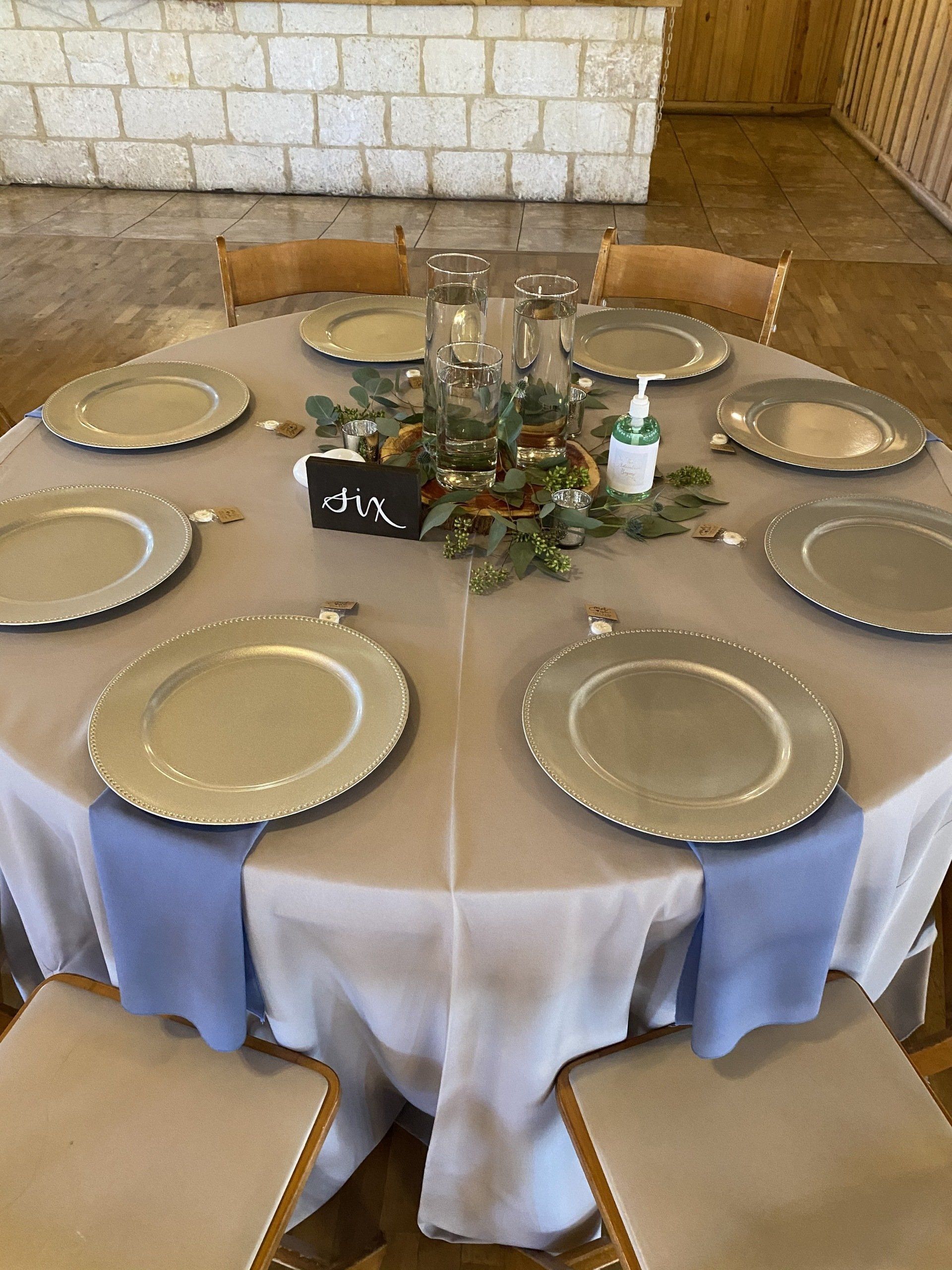 A round table set for a wedding, featuring silver chargers, light blue napkins, and a floral centerpiece labeled 
