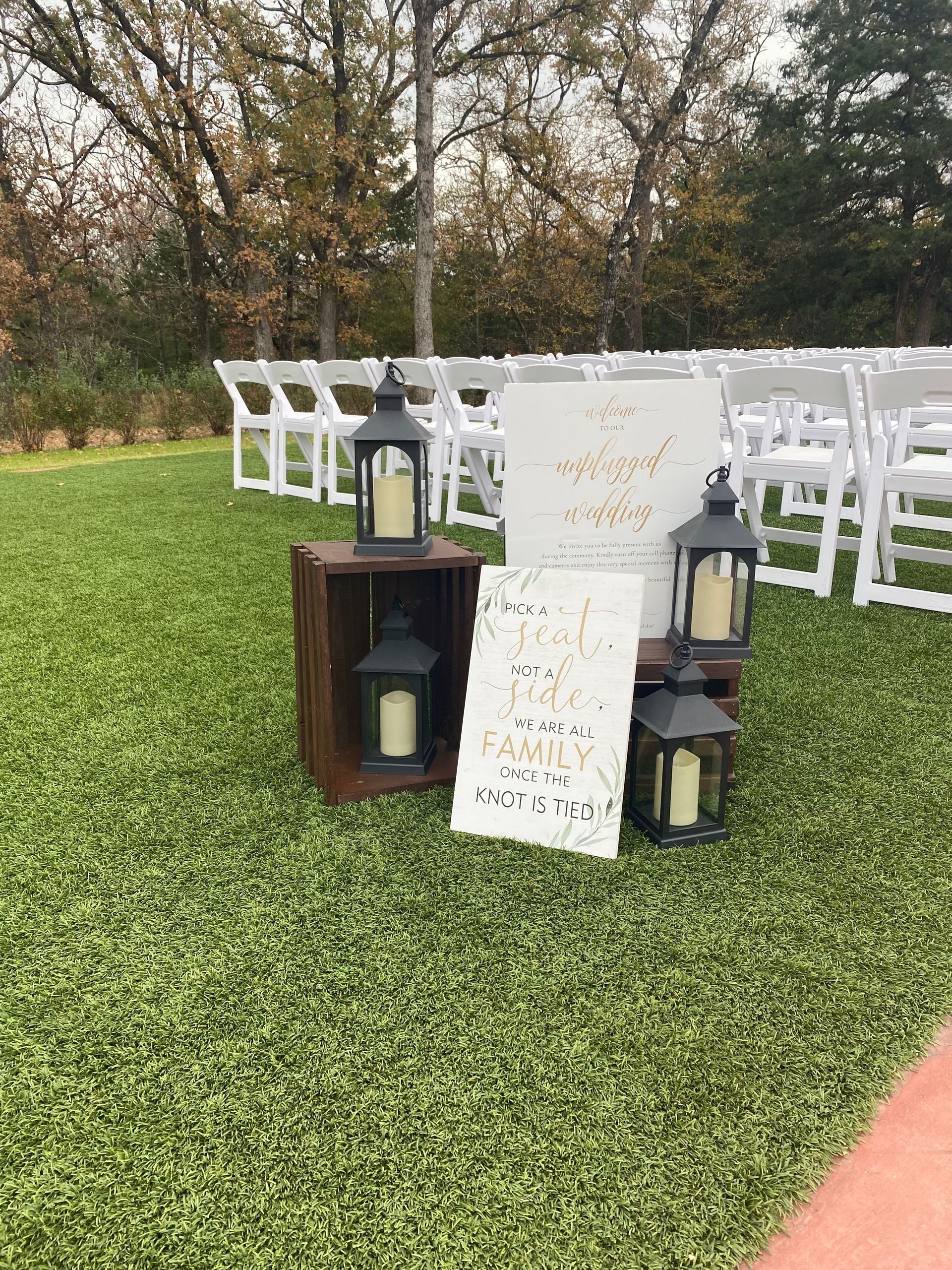 A wedding ceremony is taking place on a lush green field with white chairs and lanterns.
