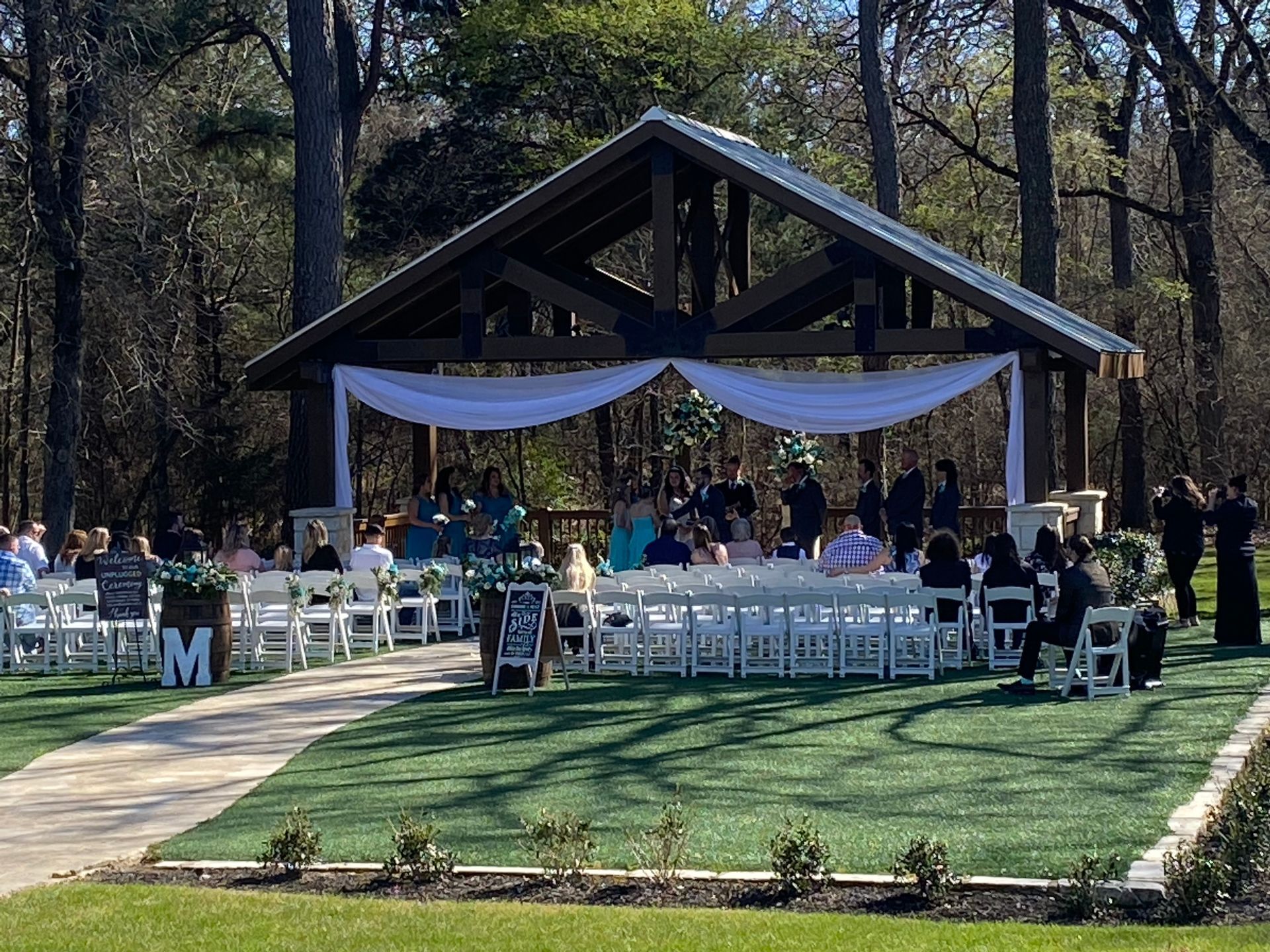 A wedding ceremony is taking place under a pavilion in the woods.