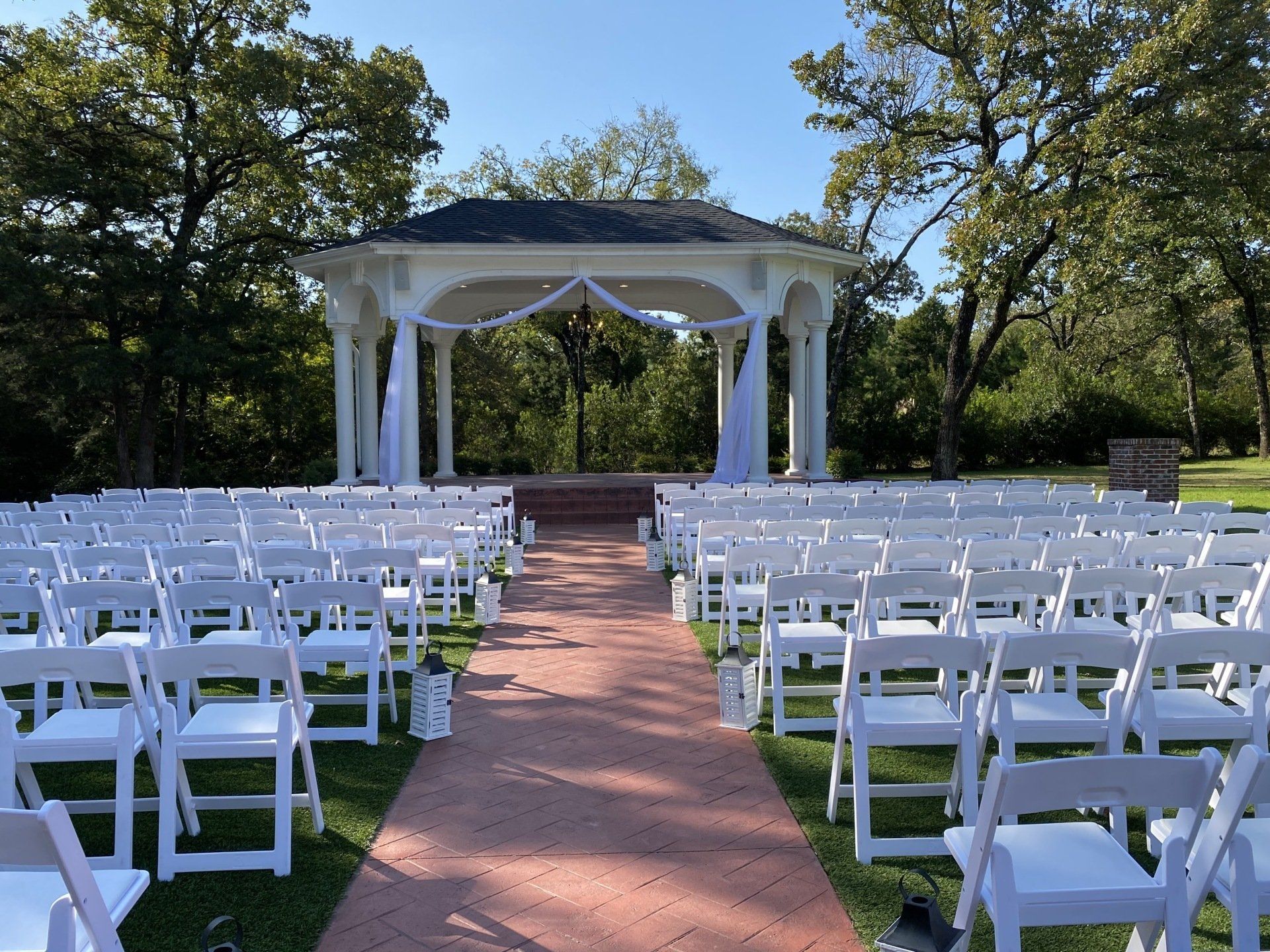 A red brick aisle leads to a white gazebo set up for an outdoor wedding with rows of white folding chairs on grassy lawns.
