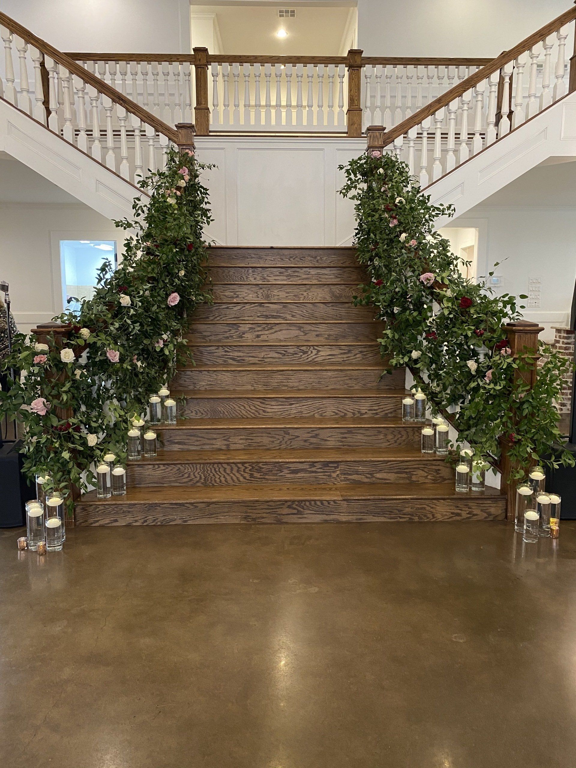 A grand wooden staircase decorated with greenery and flowers, accented by candle-filled vases lining the base.