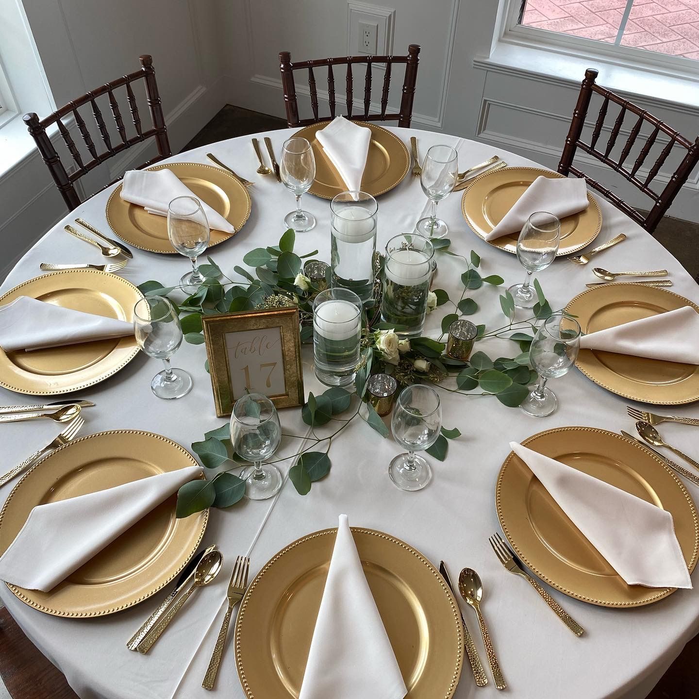 A round table set for an event features gold charger plates, white napkins, wine glasses, and a center floral arrangement.