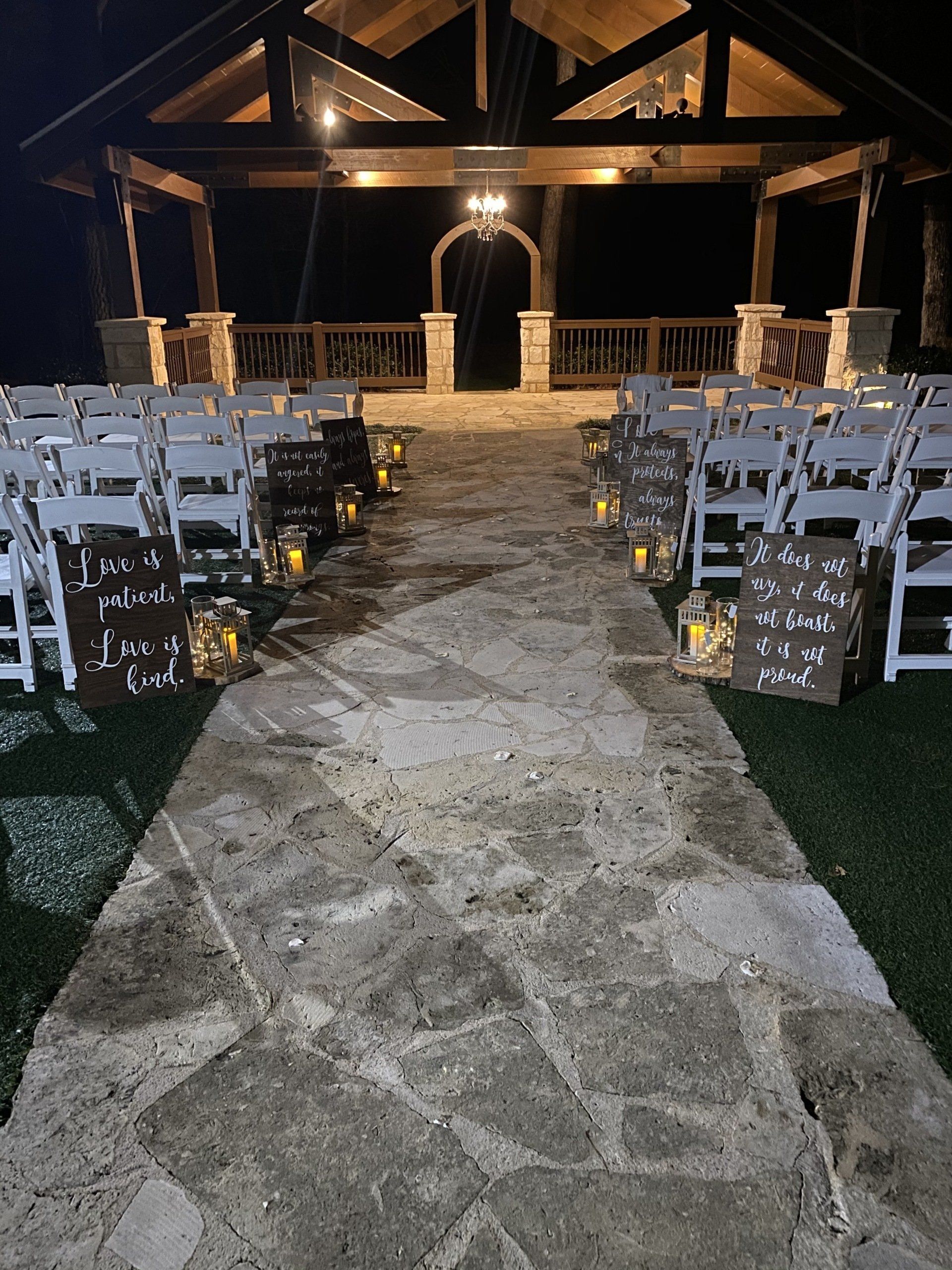 An outdoor wedding ceremony aisle lined with white chairs, lanterns, and signs at night leading to a rustic wood pavilion.