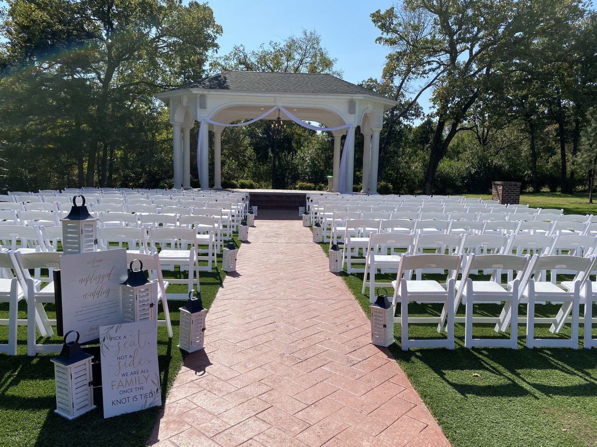 An outdoor wedding venue with rows of white folding chairs facing a white gazebo on a sunny day.