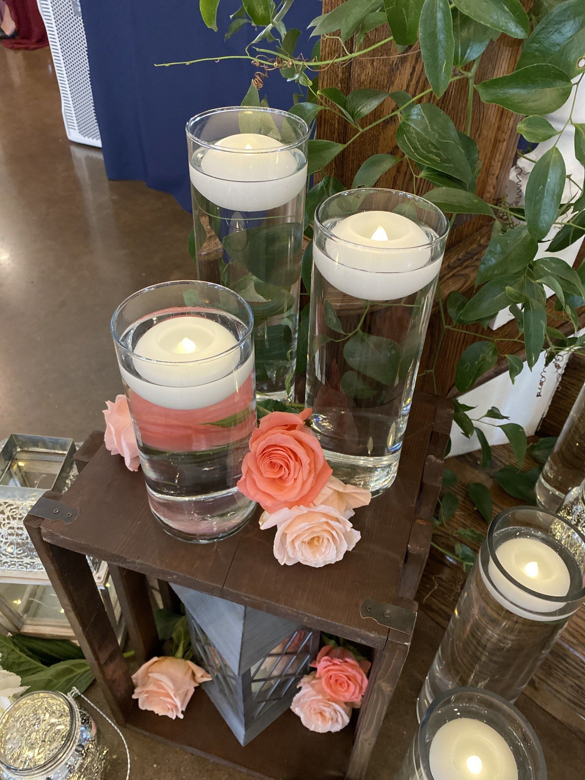Floating candles in clear glass cylinders, accented with peach roses on a rustic wooden stand.