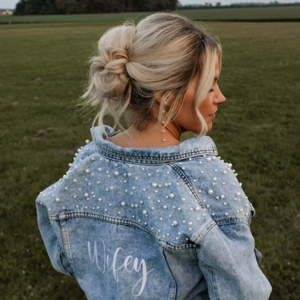 Blonde woman in pearl-studded denim jacket, seen from behind in a grassy field, hair in a loose bun.