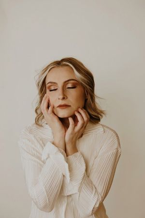Woman in a cream sweater with eyes closed, gently touching her face against a plain beige background