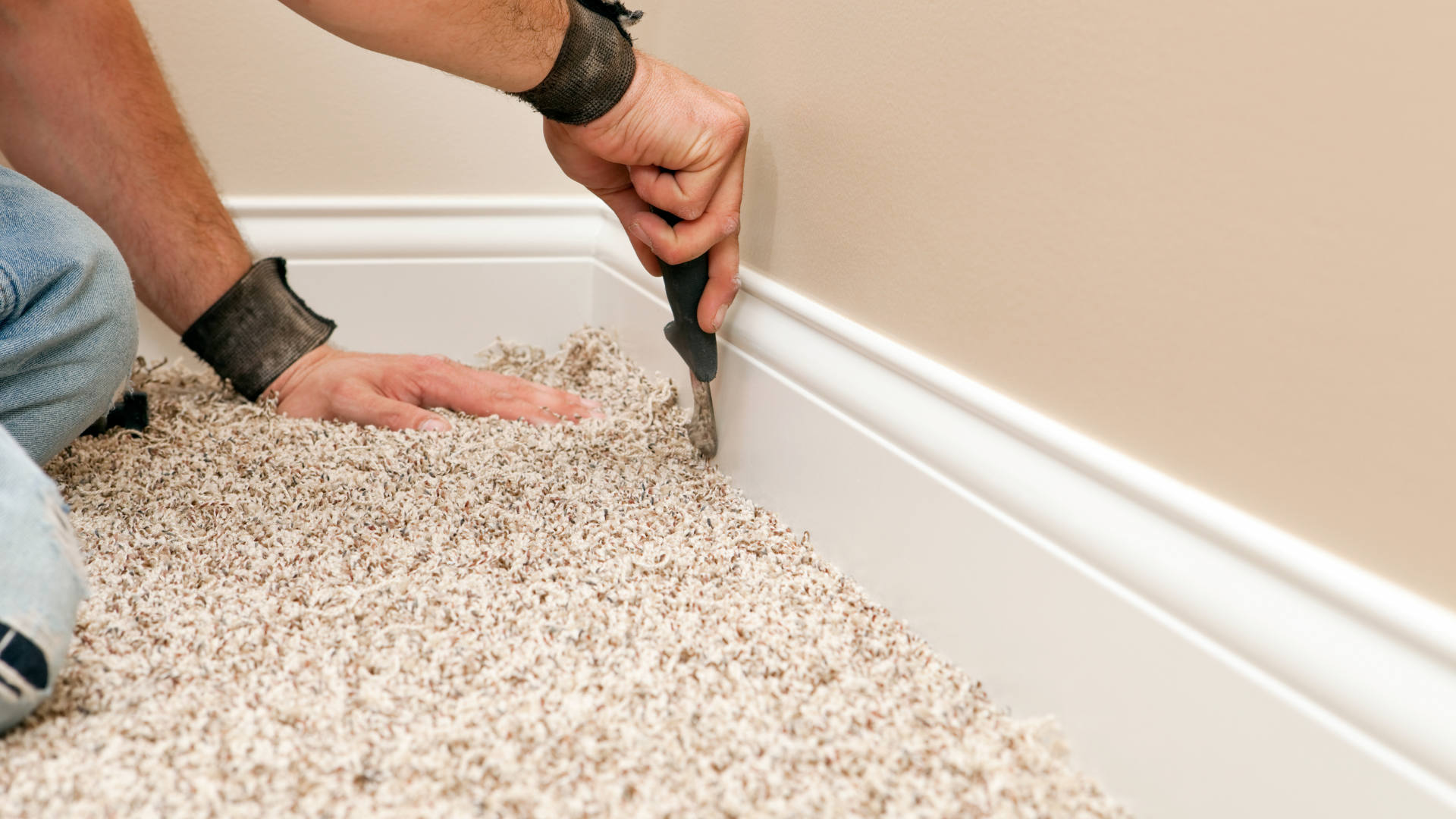 Person using a utility knife to trim carpet along a white baseboard in a room with beige walls.