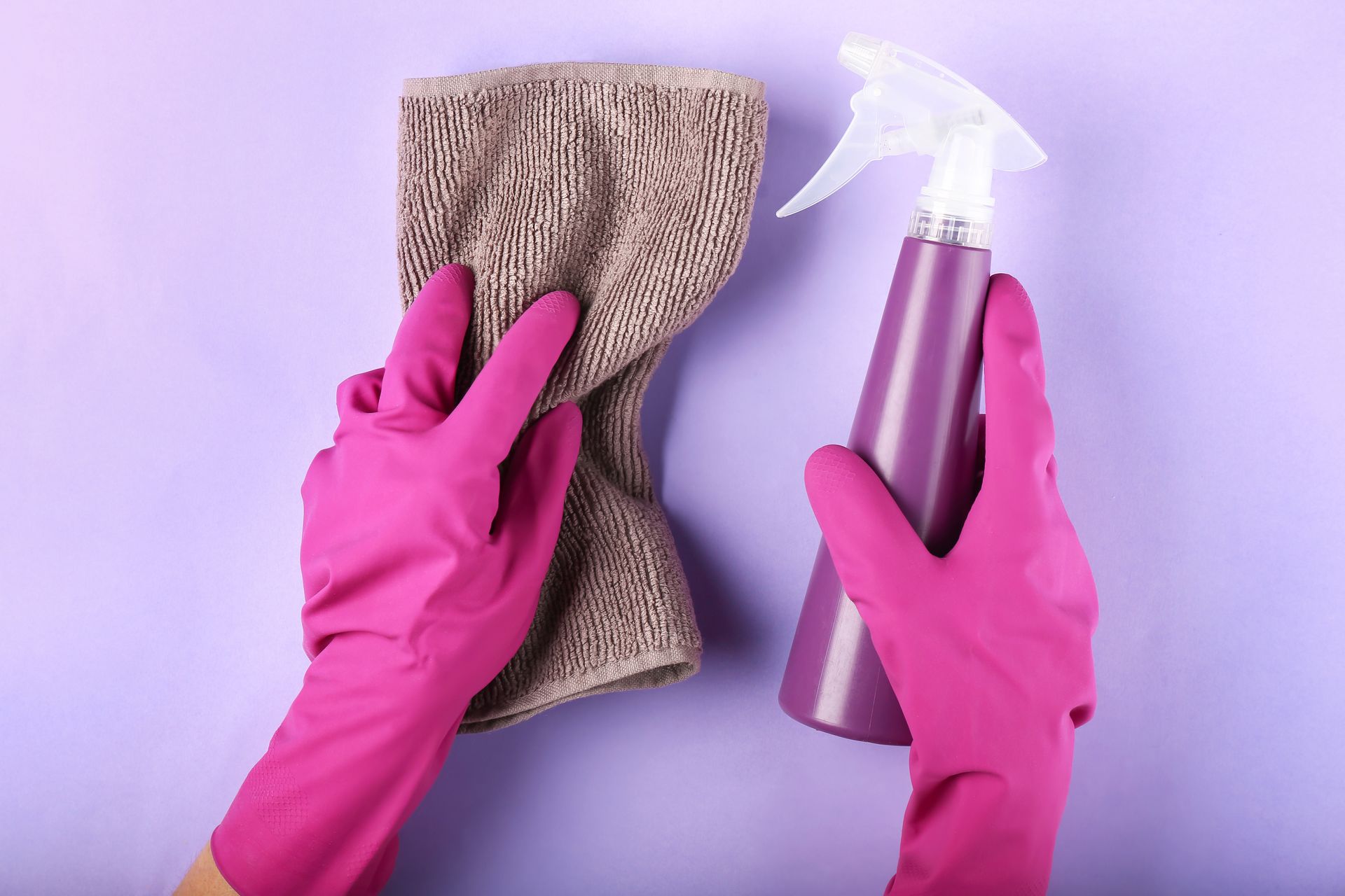 Hands in pink gloves hold a cleaning spray bottle and cloth against a purple wall.