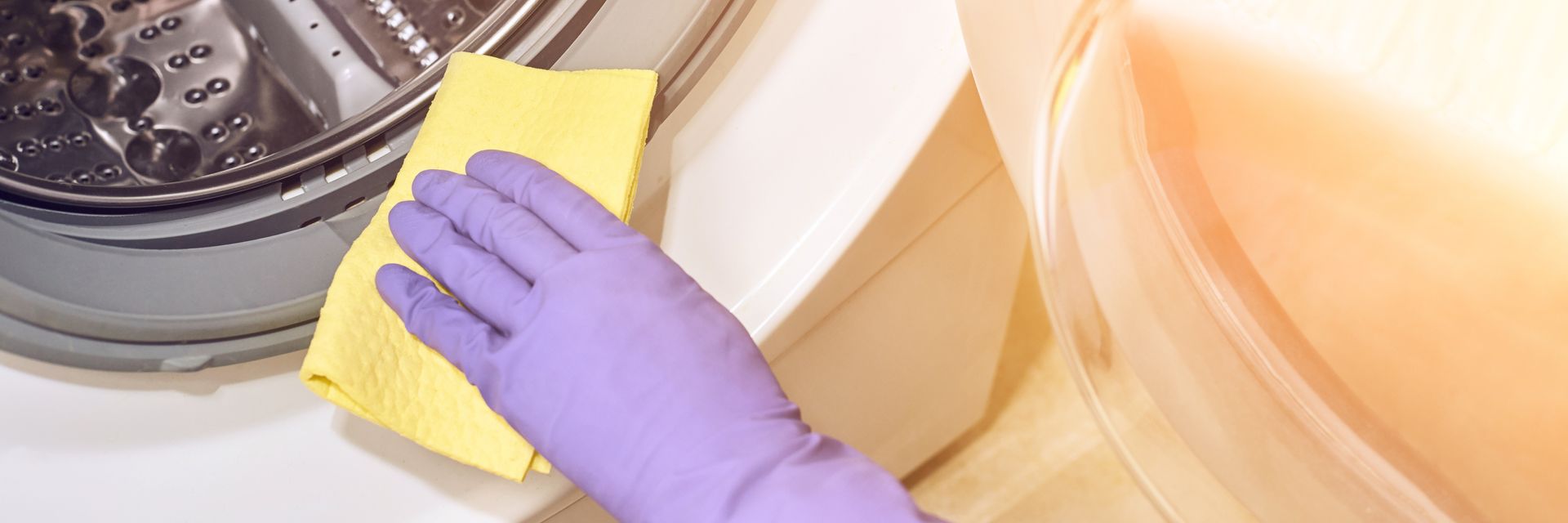A person wearing purple gloves cleans a washing machine with a yellow cloth.