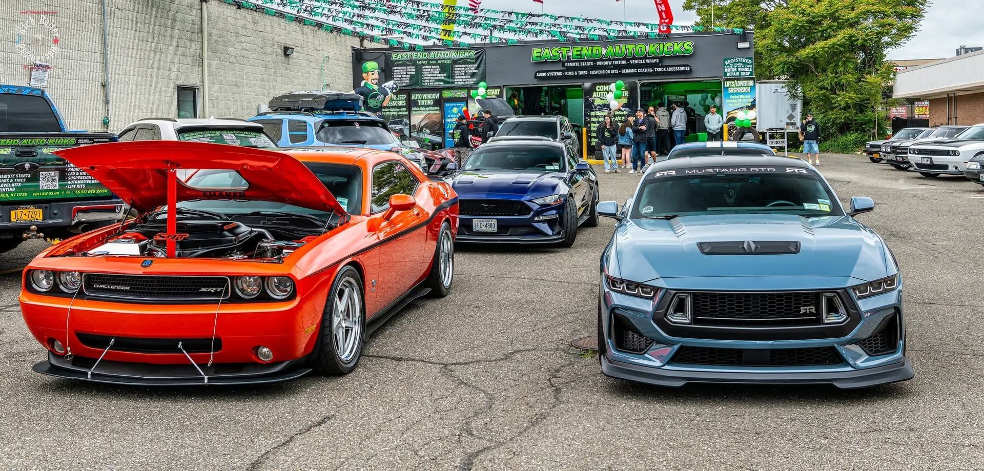 Classic muscle cars lined up outdoors, including an orange car with its hood open and a blue car in front. | East End Auto Kicks