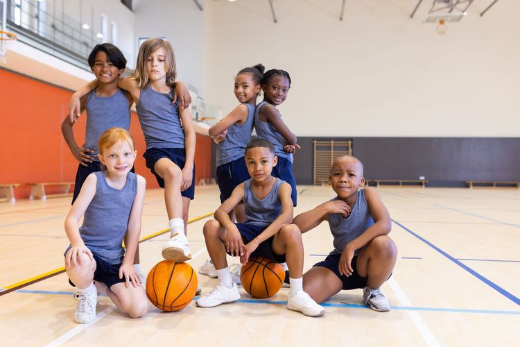 Children posing with basketballs in a gym, smiling on a court with orange cones and a hoop in the background