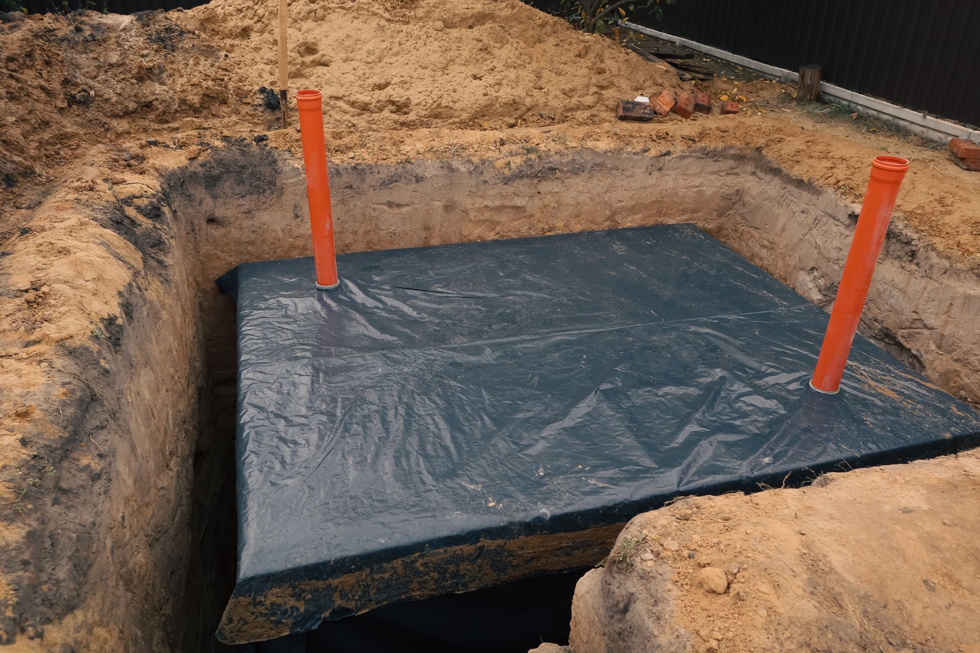 Black plastic liner in an excavated pit with two orange stakes in sandy soil