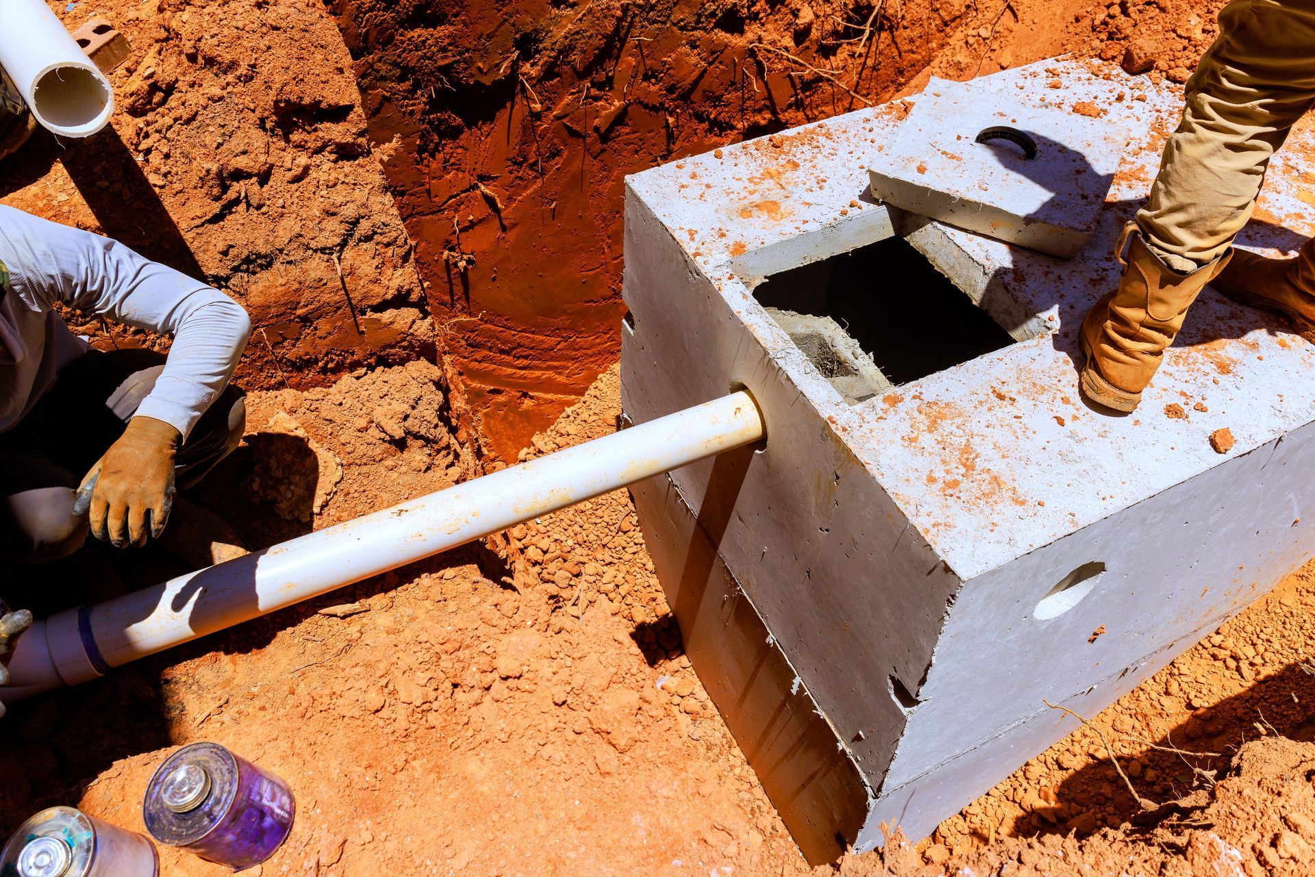 Workers installing a white pipe into a concrete utility box in red soil at a construction site
