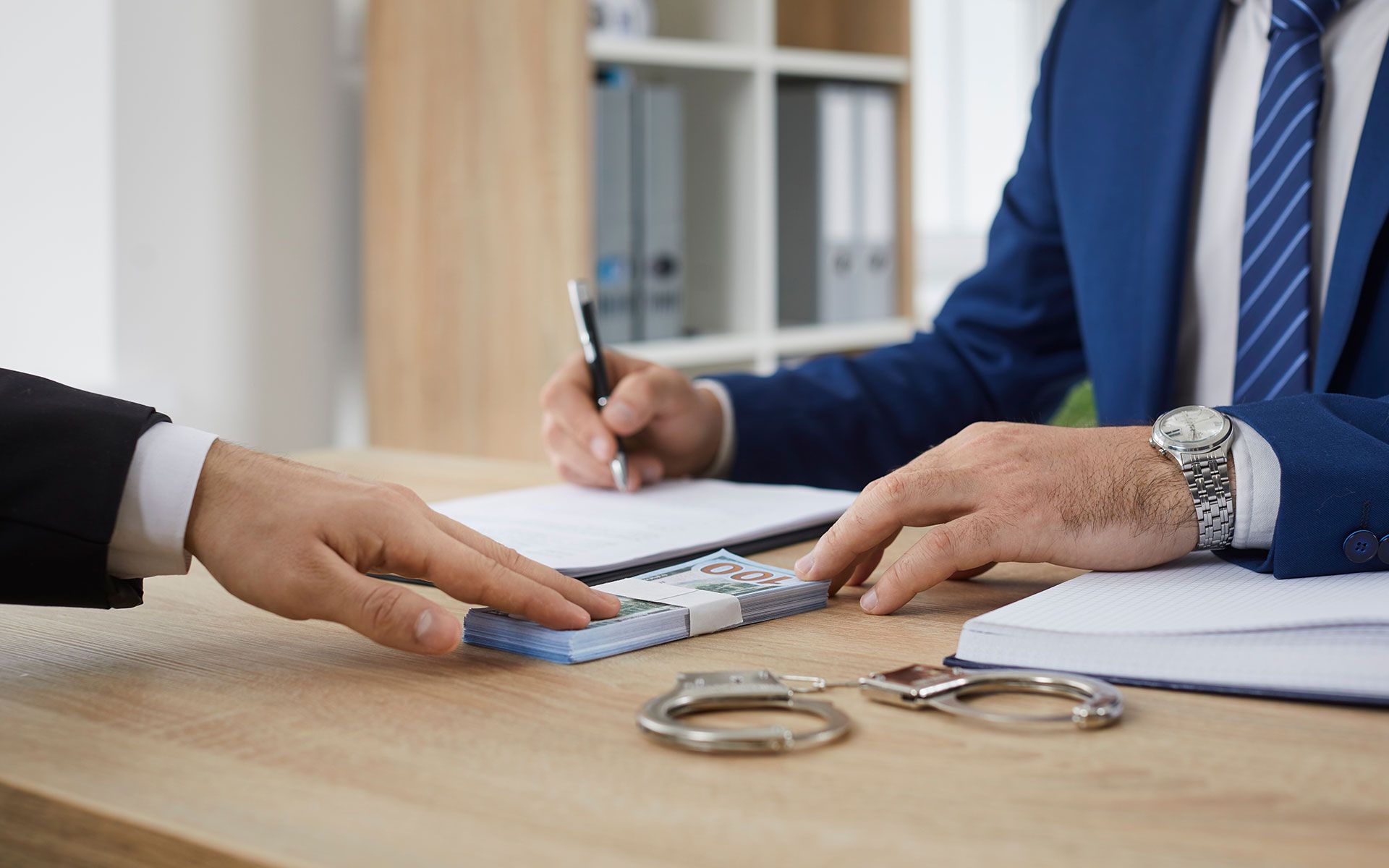 A person hands cash to another at a desk with handcuffs. One person signs a document.